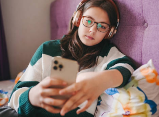 A teenage girl wearing glasses, headphones, and a green-and-white striped sweater lies back against a purple upholstered headboard while holding and looking intently at her smartphone.