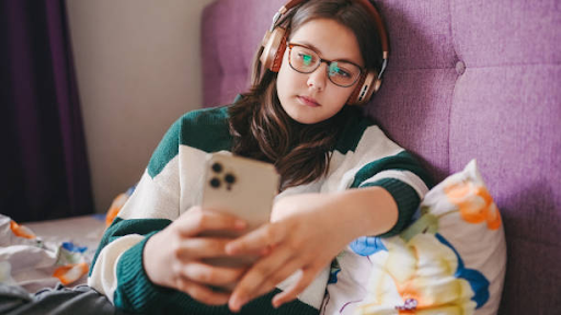 Young Person Wearing Headphones and Glasses Looking at Phone A teenage girl wearing glasses, headphones, and a green-and-white striped sweater lies back against a purple upholstered headboard while holding and looking intently at her smartphone.