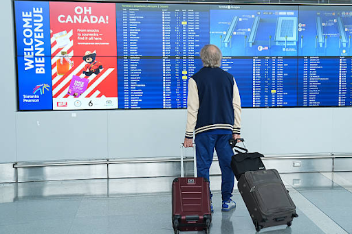 Traveler Checking Flight Departure Board at Toronto Pearson Airport A traveler seen from behind holding two rolling suitcases while standing in front of a large digital flight information board at Toronto Pearson Airport. The display shows flight schedules alongside a red advertisement featuring the text "OH CANADA!"