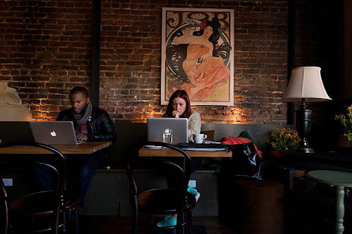 Remote Workers Seated at Tables in a Cozy Cafe A man and a woman sitting side-by-side at separate wooden tables in a dimly lit, cozy coffee shop. Both are focused on open laptops against an exposed red brick wall adorned with a framed art print and a classic table lamp.